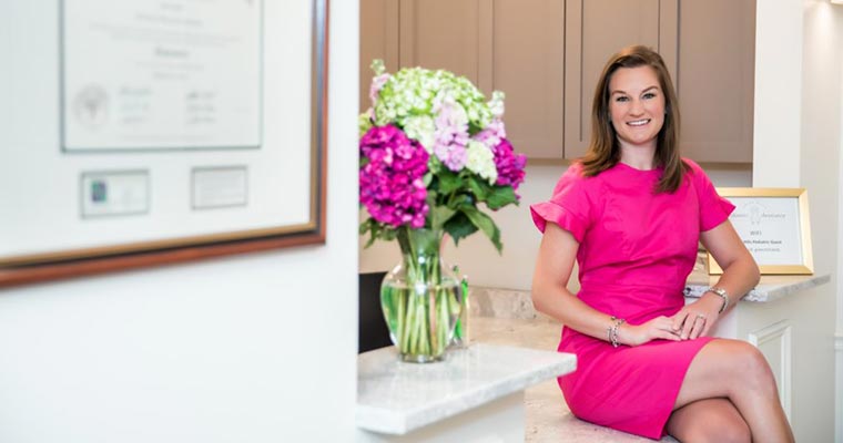 Dr. Brittany Adamiak in a bright pink dress sitting on the front desk counter smiling, ready for reopening.